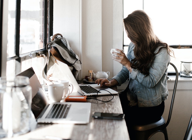 Lady in a grey and blue jacket holds coffee while working on a laptop